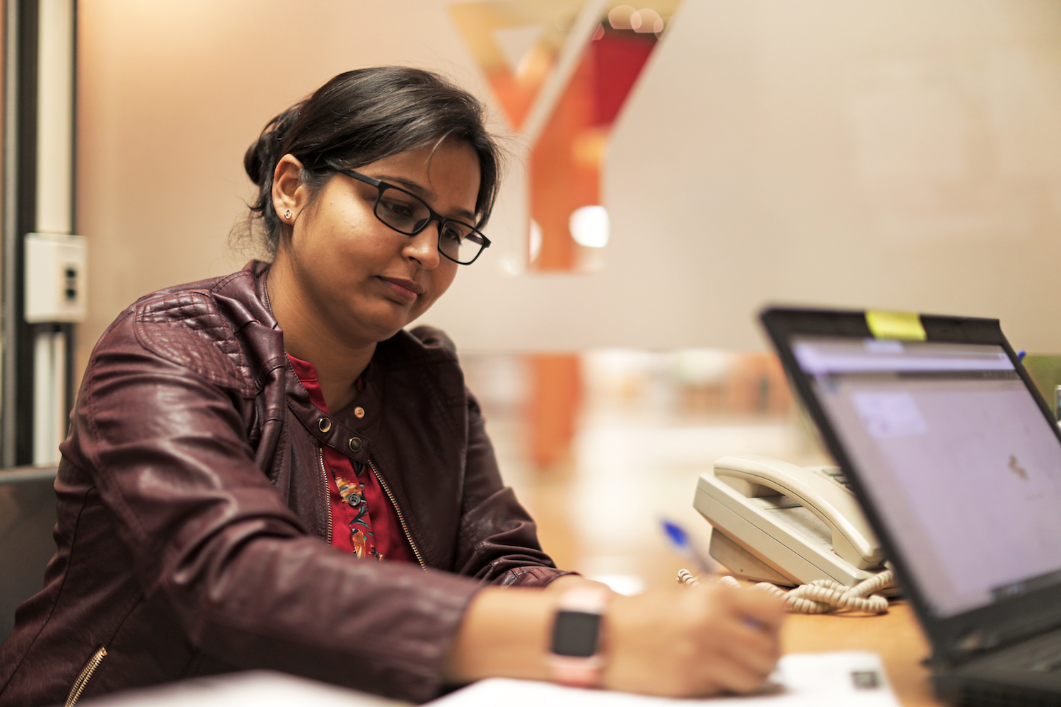 A woman in a purple leather jacket and glasses fills out a form at YMCA Settlement Services.